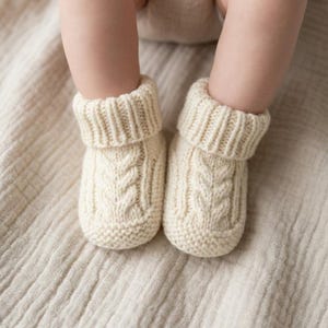 May include: A pair of cream-colored knitted baby booties featuring a vertical cable knit pattern on the front, a textured garter stitch base, and a thick ribbed cuff at the top. The booties are crafted from a soft yarn and have a folded-over collar.