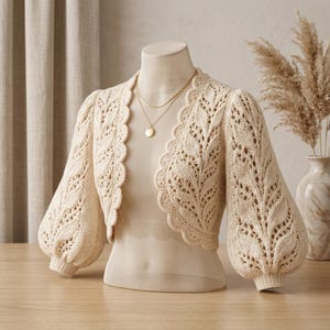 Può includere: Un bolero in maglia color crema caratterizzato da un intricato motivo traforato a foglie, maniche a sbuffo voluminose con polsini a coste e un bordo anteriore smerlato. Il capo è esposto su un manichino con una semplice collana con ciondolo dorato.