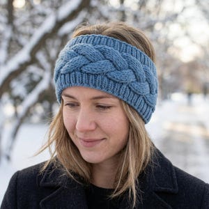 May include: A dusty blue knitted headband featuring a prominent central cable knit braid pattern. The accessory is constructed with a ribbed knit texture along the top and bottom edges, providing a snug fit around the head with a thick yarn texture.