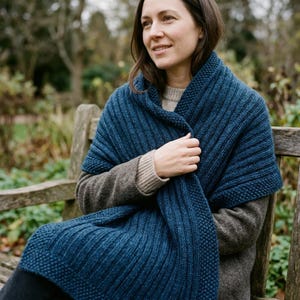 May include: A deep blue knitted shawl featuring a textured ribbed pattern throughout the main body and a seed stitch border along the edges. The garment is draped over a neutral-toned long-sleeved sweater with ribbed cuffs.