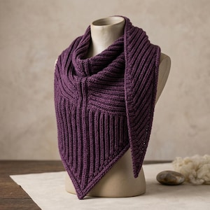 Puede incluir: Un pañuelo triangular de color morado intenso con un patrón de punto acanalado vertical prominente. El pañuelo está drapeado alrededor del cuello de un maniquí, mostrando su construcción texturizada y su dobladillo en punta.
