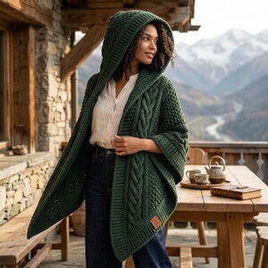 Könnte beinhalten: Eine dunkelwaldgrüne Kapuzenstrickjacke mit grobem Zopfmuster, breitem Rippstrickrand und einem kleinen rechteckigen Etikett in Lederoptik an der unteren Ecke. Das Kleidungsstück wird über einem hellen Hemd und einer dunklen Jeans getragen.