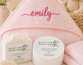 May include: A pink hooded baby towel embroidered with the name "emily" in cursive script. The image also shows a bar of soap, a jar of baby body butter, a wooden brush, and a stuffed bunny.