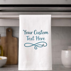 May include: White kitchen towel with the text "Your Custom Text Here" in teal script and a decorative flourish. The towel is hanging on a stainless steel oven handle. A wooden chopping board is visible in the background.