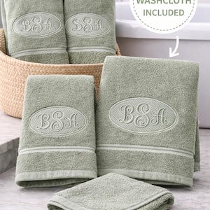 May include: Sage green bath towels and washcloths with embroidered initials. The towels are displayed in a woven basket and on a tiled surface. A free washcloth is included. The towels have a decorative border.