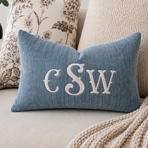 May include: A blue decorative pillow with the embroidered initials "CSW" in white. The pillow is rectangular and has a textured appearance. It is placed on a light-colored sofa, with other pillows in the background.