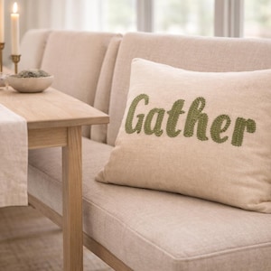 Peut inclure: Un coussin beige avec le mot "Gather" brodé en vert. Le coussin est posé sur un banc beige rembourré. Une table en bois avec un chemin de table en lin et des bougies est à l'arrière-plan.