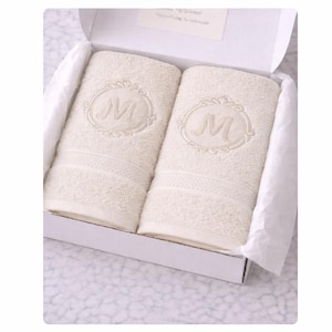 Peut inclure: Deux serviettes en coton couleur crème pliées dans une boîte cadeau blanche. Chaque serviette présente une lettre M brodée au centre d'un cadre circulaire décoratif. Les serviettes ont une bordure texturée et sont placées dans du papier de soie.