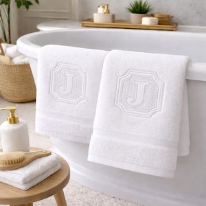 Puede incluir: Dos toallas de baño blancas con la letra "J" bordada en un marco decorativo. Las toallas cuelgan del borde de una bañera blanca. Un pequeño taburete de madera sostiene toallas dobladas y un cepillo.