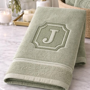 Pode incluir: Toalha de banho verde sálvia com uma letra "J" bordada em uma moldura decorativa. A toalha está dobrada e colocada sobre uma superfície de mármore. Outras toalhas são visíveis em uma cesta de vime ao fundo.