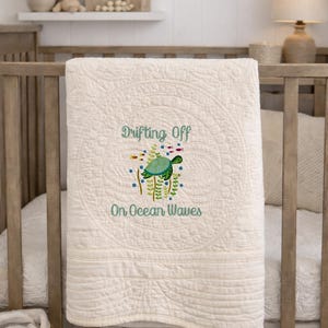 May include: Cream-colored baby quilt with an embroidered sea turtle design. The quilt features the text "Drifting Off On Ocean Waves". The quilt is draped over a wooden crib.