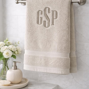 Op de afbeelding: Een beige monogram badhanddoek met de letters "GSP" geborduurd in een bijpassende kleur. De handdoek hangt aan een zilveren handdoekrek in een badkamer. De badkamer heeft witte tegels en een marmeren aanrecht.