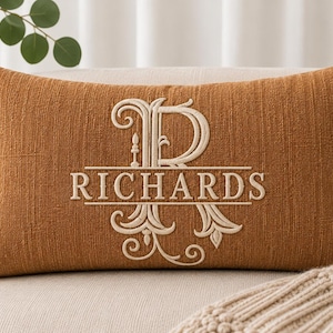 May include: A rectangular burnt orange throw pillow featuring a large, cream-coloured embroidered monogram letter R with the name RICHARDS stitched horizontally across the centre in a serif font, set against a textured fabric surface.