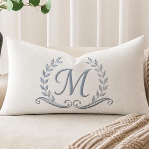 Peut inclure: Un coussin rectangulaire blanc cassé avec une lettre monogramme M brodée au centre en fil bleu cendré. La lettre est encadrée par un motif symétrique de couronne de laurier avec des volutes décoratives à la base, cousues en bleu.