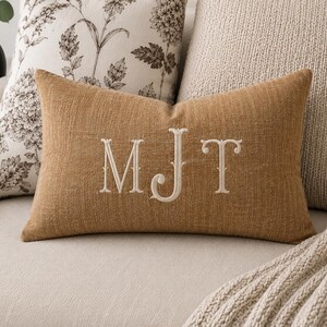 May include: A rectangular throw pillow in a textured mustard-brown fabric featuring the embroidered white monogram letters MJT centered on the front. The pillow has a clean, finished edge and a visible weave pattern across the surface.