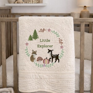 Puede incluir: Edredón de bebé color crema con un diseño bordado de bosque. El edredón presenta una corona circular con un zorro, un ciervo, setas y el texto "Little Explorer". El edredón está sobre una cuna de madera.