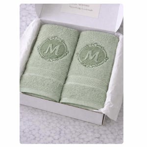 May include: Two sage green cotton towels folded neatly inside a white cardboard gift box. Each towel features a centered, circular embroidered emblem with a decorative border surrounding the capital letter M in a matching sage green thread.