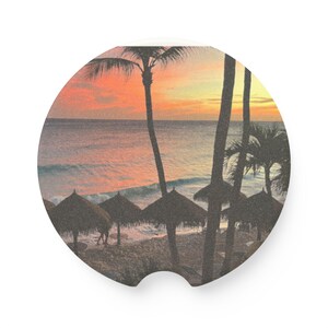 May include: Circular car coaster featuring a beach scene at sunset. The image shows palm trees, thatched umbrellas, and waves under a vibrant orange and yellow sky. The design evokes a tropical vacation vibe.