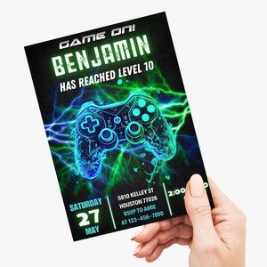 May include: A green and blue video game controller with lightning bolts on a black background. The text "GAME ON! BENJAMIN HAS REACHED LEVEL 10" is at the top of the invitation. The text "SATURDAY 27 MAY" is in the bottom left corner. The text "5810 KELLEY ST HOUSTON 77026 RSVP TO AMIE AT 123-456-7890 2:00" is in the bottom right corner.