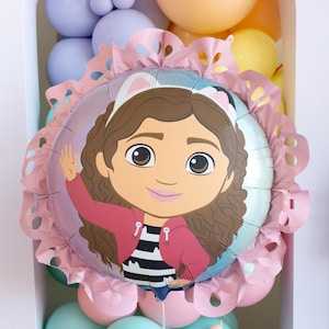 May include: A round foil balloon featuring a cartoon girl with brown hair, wearing a pink jacket and a black and white striped shirt. The balloon is surrounded by a pink floral-shaped border and rests on a cluster of pastel-colored balloons.