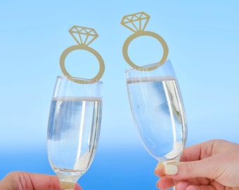 May include: Two champagne flutes clink together, each adorned with a gold ring-shaped charm featuring a diamond design. The glasses are filled with a bubbly liquid, set against a bright blue sky background.