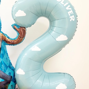 Puede incluir: Globo azul claro con forma de n&uacute;mero 2, decorado con nubes blancas y el nombre "OLIVER". Forma parte de un arreglo m&aacute;s grande con otros globos de colores y un personaje de dibujos animados.