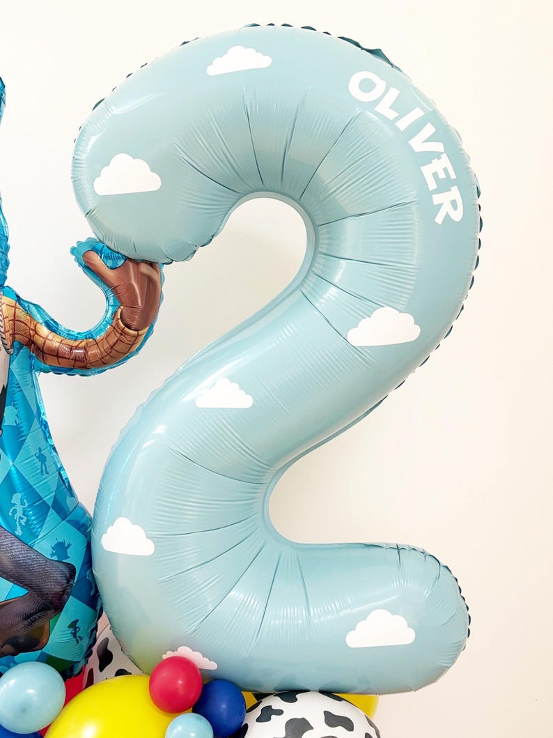 Puede incluir: Globo azul claro con forma de n&uacute;mero 2, decorado con nubes blancas y el nombre "OLIVER". Forma parte de un arreglo m&aacute;s grande con otros globos de colores y un personaje de dibujos animados.