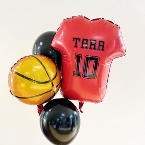 Peut inclure: Un ensemble de ballons, dont un ballon rouge en forme de maillot avec le nom "TARA" et le numéro "10" en noir. Un ballon jaune en forme de ballon de basket et deux ballons ronds noirs complètent l'ensemble. Des ficelles blanches sont visibles.