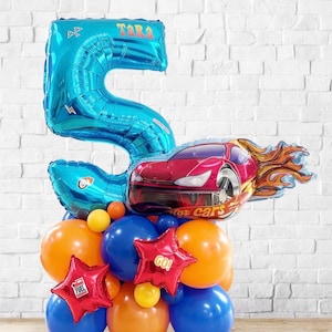 May include: A celebratory balloon display. It features a large blue number 5 balloon with the name "Tara", a red race car balloon with flames, and a cluster of orange, blue, and yellow balloons. Red star-shaped balloons with "GO!" and "FUEL" text are also included.