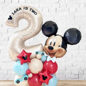 May include: A festive balloon arrangement featuring a large number two balloon with the text "TARA IS TWO". The arrangement includes a Mickey Mouse balloon, red star balloons, and various other balloons in red, white, and blue.