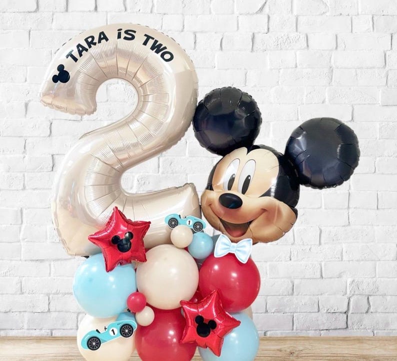 May include: A festive balloon arrangement featuring a large number two balloon with the text "TARA IS TWO". The arrangement includes a Mickey Mouse balloon, red star balloons, and various other balloons in red, white, and blue.