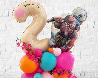 May include: A festive balloon arrangement for a second birthday. The arrangement features a large number "2" balloon with the text "Tara is two", a Moana-themed balloon, and a cluster of colorful balloons in pink, orange, and teal.