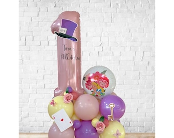 May include: A whimsical balloon arrangement for a first birthday. It features a large pink number one balloon with a Mad Hatter hat, surrounded by smaller balloons in pastel colors, a playing card, and a key.