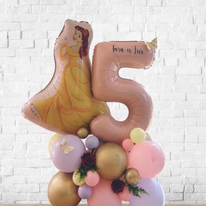 Puede incluir: Arreglo festivo de globos con un gran globo con el número 5 y un globo con forma de Bella. El arreglo incluye globos dorados, rosas y morados, con el texto "Tara is Five". Mariposas y flores decorativas.