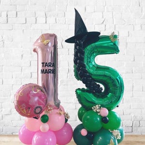 以下が含まれることがあります： 誕生日のお祝い用の2つのバルーンアレンジメント。1つは「Tara Marie」という名前と丸いピンクの風船が付いたピンク色の数字の1です。もう1つは、魔女の帽子と黒い髪の緑色の数字の5です。
