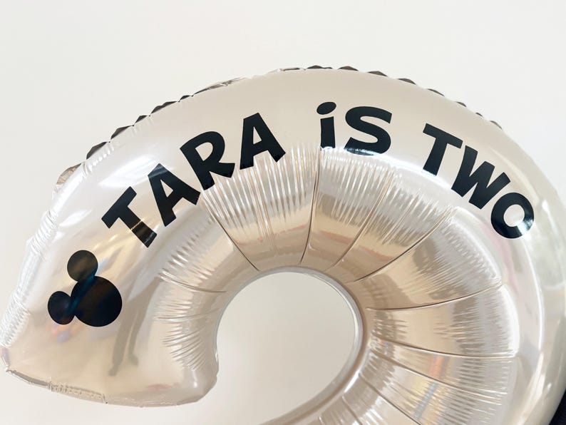 May include: Silver foil number two balloon with the text "TARA is TWO" in black. A black Mickey Mouse head is on the left side of the balloon. Perfect for a second birthday party.