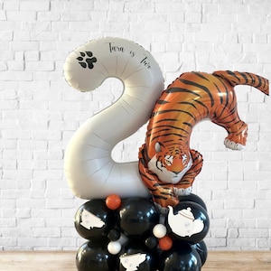 Puede incluir: Un arreglo de globos para una celebración. Un gran globo blanco con el número "2" y el texto "Tara is Two" y una huella de pata. Un globo con forma de tigre se encuentra encima de un grupo de globos negros, decorados con detalles blancos y naranjas.