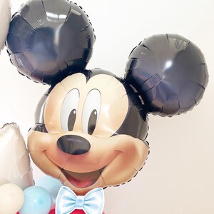 May include: A Mickey Mouse-shaped balloon with black ears and a smiling face. The balloon has a light blue bow tie and is part of a larger balloon arrangement with red, white, and blue balloons. Perfect for a birthday party.