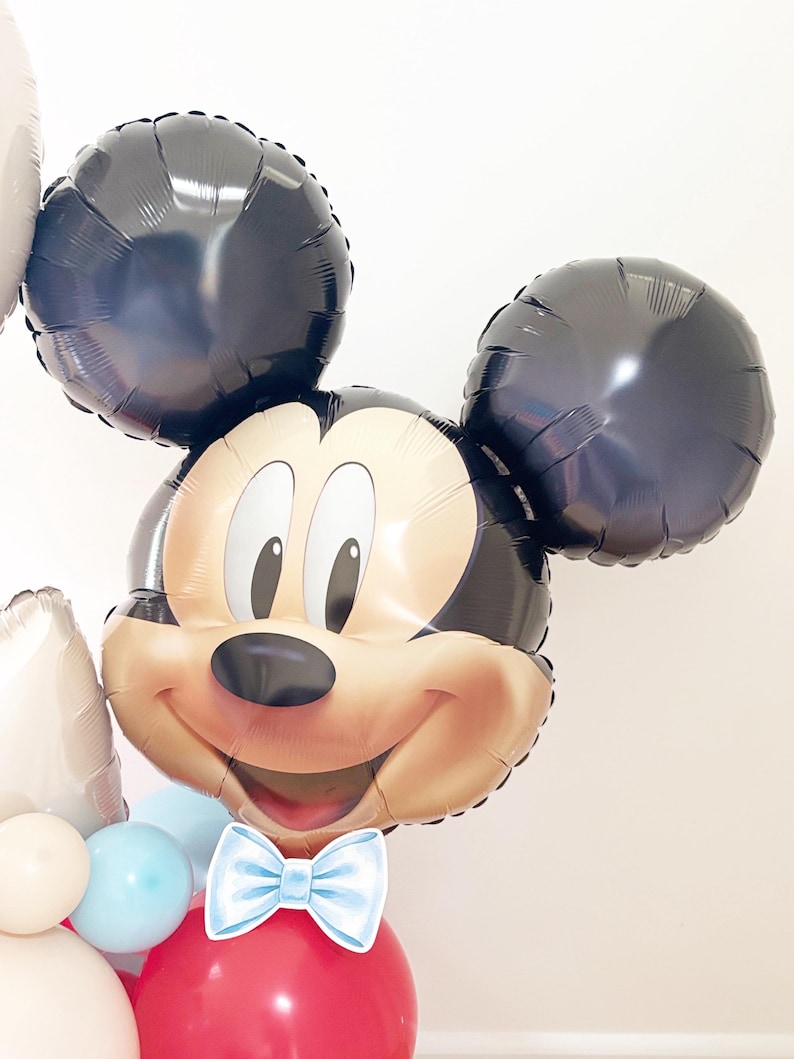 May include: A Mickey Mouse-shaped balloon with black ears and a smiling face. The balloon has a light blue bow tie and is part of a larger balloon arrangement with red, white, and blue balloons. Perfect for a birthday party.