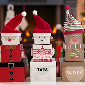 May include: Three Christmas-themed gift boxes stacked. The top boxes are decorated as Santa Claus, a bear, and a reindeer, each with a matching hat. The boxes are red, white, and brown, with the names "ELINA", "TARA", and "OLIVER" on them.