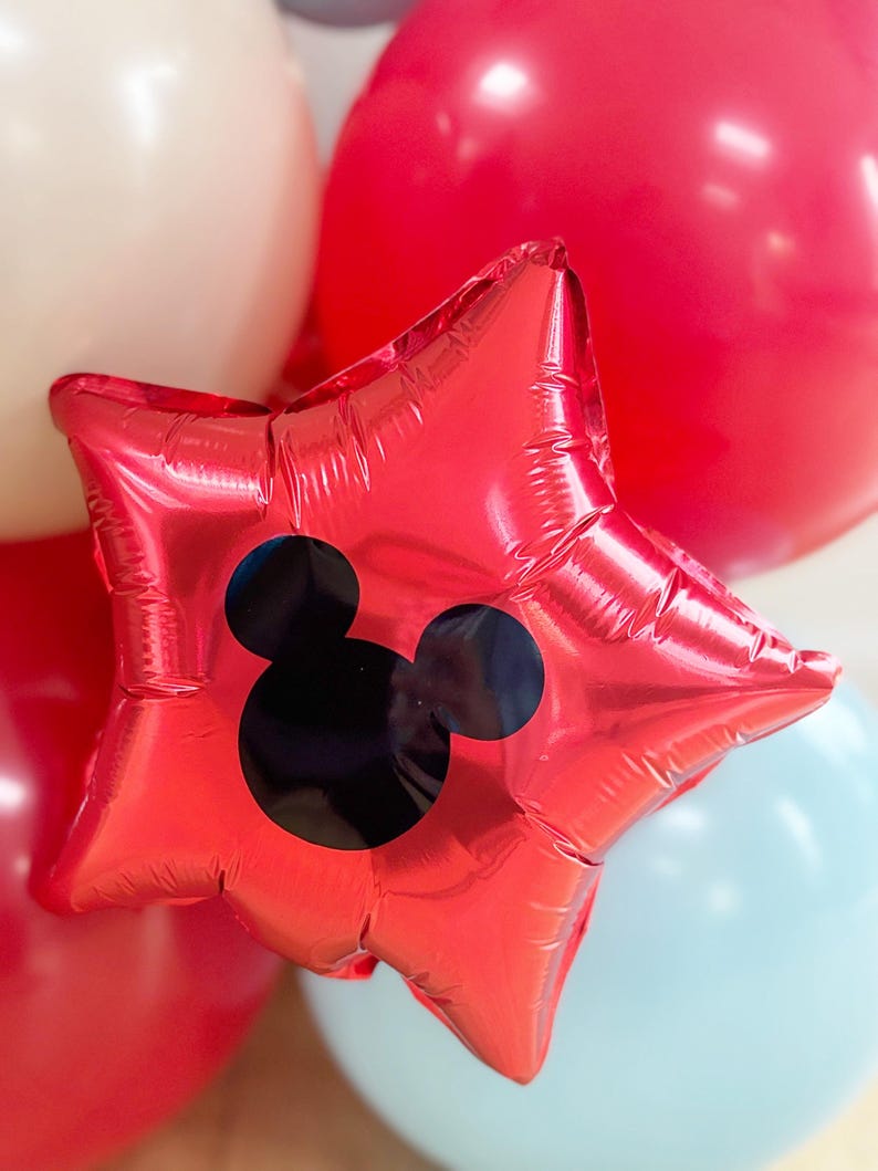 May include: A red star-shaped balloon with a black Mickey Mouse silhouette. The balloon is surrounded by other balloons in red, white, and light blue. This festive balloon is perfect for a birthday party or other celebration.
