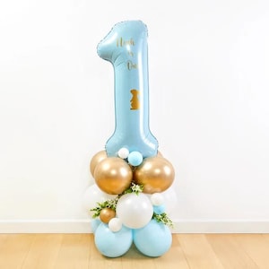 May include: A light blue number one balloon with gold text that reads "Noah is One." The balloon is part of a balloon arrangement with gold, white, and light blue balloons. The arrangement is decorated with small white flowers.