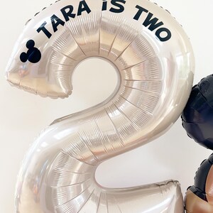 May include: A large silver number two balloon with the text "TARA is TWO" in black. A Mickey Mouse silhouette is on the left side of the balloon. Other balloons in red, black, and white are visible in the background. Party decorations.
