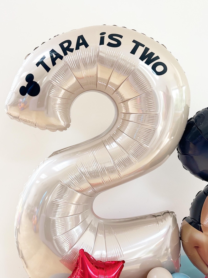 May include: A large silver number two balloon with the text "TARA is TWO" in black. A Mickey Mouse silhouette is on the left side of the balloon. Other balloons in red, black, and white are visible in the background. Party decorations.
