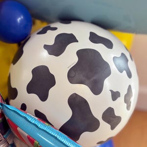 Puede incluir: Un gran globo blanco con manchas negras de vaca es el punto focal de esta imagen. Tambi&eacute;n son visibles otros globos en azul, amarillo y un globo de aluminio con rayas rojas y blancas. Los globos son probablemente para una fiesta o celebraci&oacute;n.