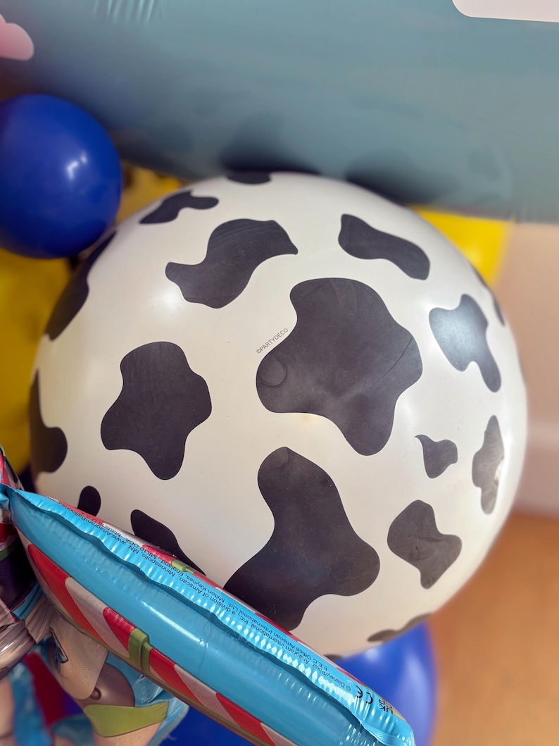 Puede incluir: Un gran globo blanco con manchas negras de vaca es el punto focal de esta imagen. Tambi&eacute;n son visibles otros globos en azul, amarillo y un globo de aluminio con rayas rojas y blancas. Los globos son probablemente para una fiesta o celebraci&oacute;n.