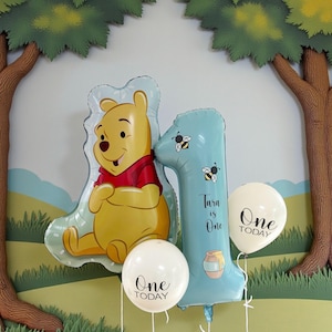 May include: A set of balloons for a first birthday. Features a Winnie the Pooh shaped balloon, a blue number one balloon decorated with bees, and two white balloons with "One Today" text.
