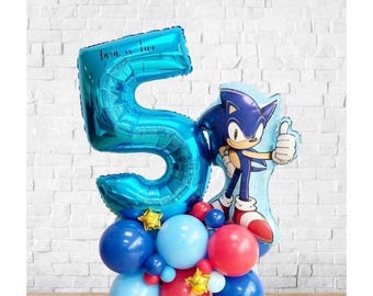 May include: A festive balloon arrangement featuring a large blue number 5 balloon with the text "Tara is two", a Sonic the Hedgehog balloon, and a cluster of blue and red balloons. Gold star-shaped balloons add a celebratory touch.