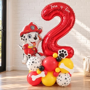 May include: A balloon arrangement featuring a large red foil number two balloon with the text Tara is Two, a Marshall Paw Patrol character foil balloon, and a cluster of red, yellow, and white balloons with black paw print patterns and bone shapes.