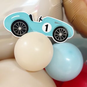 May include: A light blue race car cutout with the number 1, placed above cream and light blue balloons. The car has black wheels and a black steering wheel. The balloons are part of a party decoration.